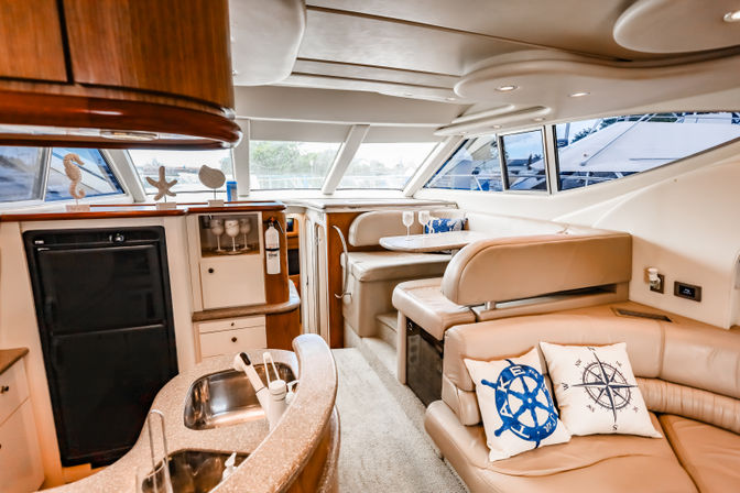 Bright luxury yacht interior with cream leather seating, compact galley sink and fridge, dinette table, nautical throw pillows and marina view through windows.