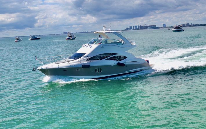 Luxury white motor yacht speeding across turquoise bay leaving a foamy wake, with other boats and a distant city skyline under a partly cloudy sky.