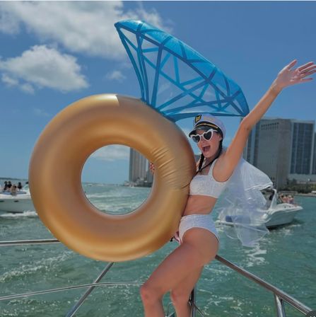 Joyful bachelorette on a yacht holding a giant gold ring float with a blue diamond topper, wearing a captain hat, white bikini, veil and heart sunglasses, posing on a sunny boat day with coastal city skyline and other vessels in the background.