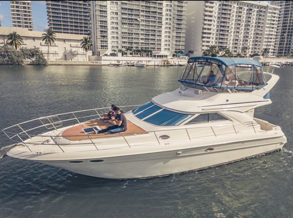 White luxury motor yacht cruising a calm urban waterway past palm‑lined waterfront high‑rise condos, two people relaxing on the bow and a person at the covered helm.