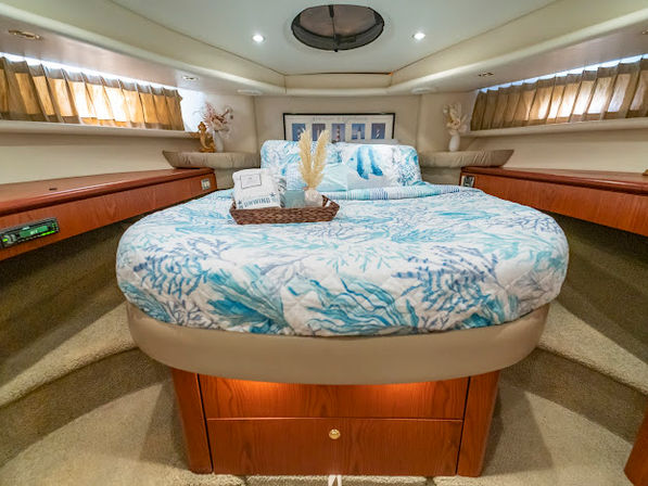 Cozy yacht master stateroom with a round bed in blue coral-print bedding, wooden storage drawers and shelves, porthole skylight, sea-themed decor, and warm ambient lighting.