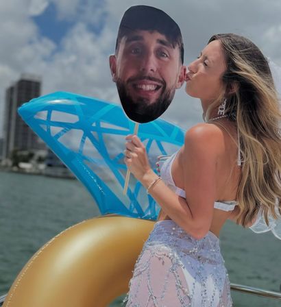 Woman in white bikini and sheer beaded cover-up on a sunny boat party kissing a large cardboard face on a stick next to a blue inflatable and gold pool float with a coastal skyline in the background