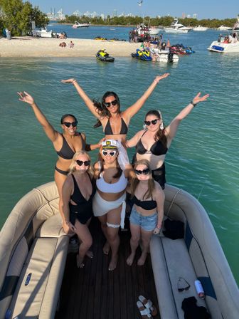 Six friends in bikinis celebrating a bachelorette on a pontoon boat in turquoise bay near a sandy sandbar with jet skis, yachts, and sunny blue skies — lively summer boat party scene.