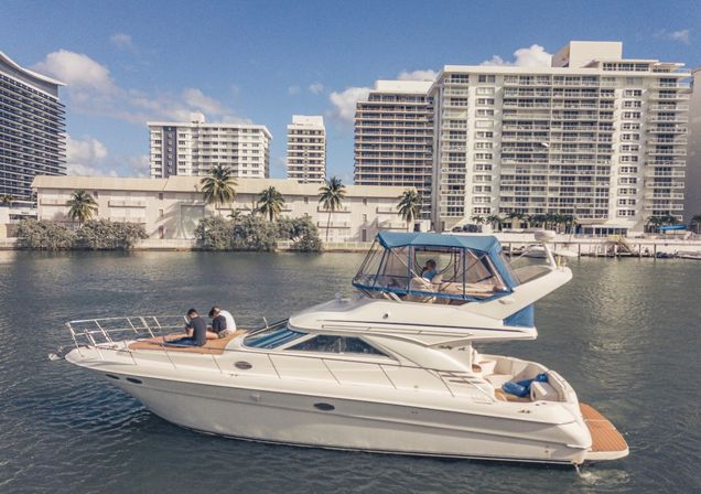 White luxury motor yacht cruising a sunny marina channel with passengers on deck, captain in the flybridge, palm trees and high-rise waterfront condos under a blue sky