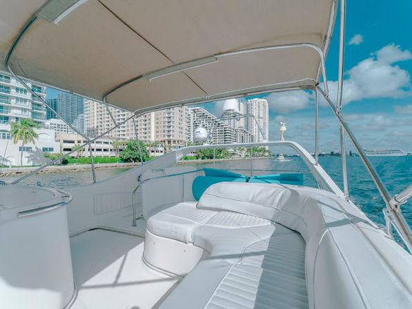 Sun-soaked luxury yacht deck with white curved seating under a beige canopy, cruising along an urban waterfront with high-rise skyline, palm trees, and blue water and sky.
