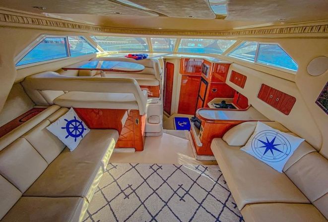 Sunlit luxury yacht salon with cream leather seating, nautical-themed throw pillows, glossy wood cabinetry, compact galley and forward windows showing marina waters.