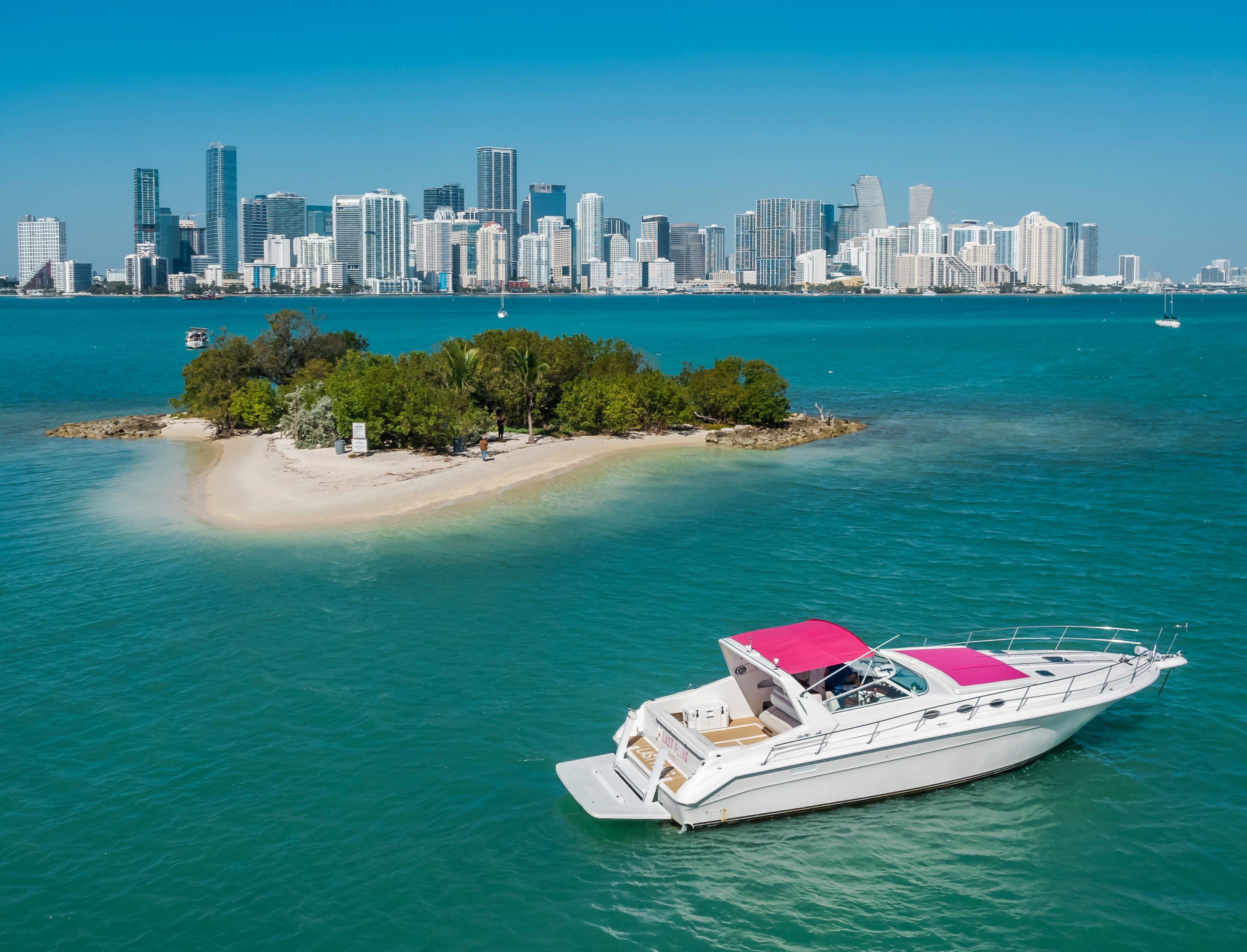 White motor yacht with pink canopy cruising turquoise waters near a small sandy island, with a modern city skyline of high‑rise buildings across the bay on a clear sunny day