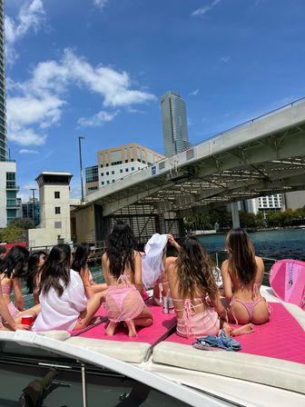 Group of women in pink swimwear lounging on a boat cruising under a drawbridge on a sunny Miami waterfront, bachelorette wearing a white veil with blue sky and city skyline in the background.