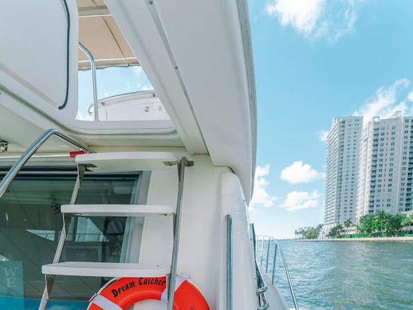 White luxury yacht stern with metal boarding ladder and orange lifebuoy on calm blue water, framed by waterfront high-rise condos and a sunny blue sky — urban marina vibe.