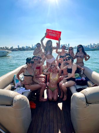 Group of friends in bikinis cheering on a pontoon boat off Miami with skyline in background, holding a red "Last Fling" sign on a sunny day