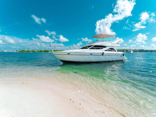 Luxury white yacht anchored near a white-sand tropical beach with turquoise water and bright blue sky dotted with clouds