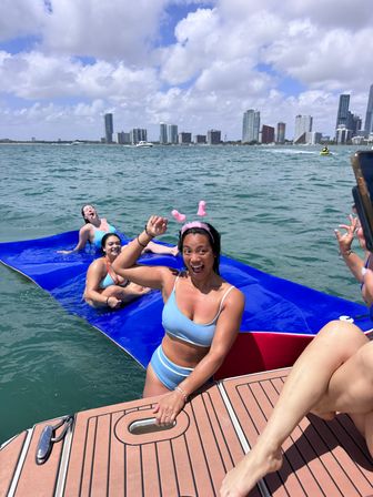 Friends laughing on a blue inflatable water mat beside a boat, woman in a light-blue bikini wearing a pink novelty headband cheering, turquoise water with the Miami skyline and partly cloudy sky in the background.