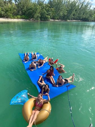 Group of friends lounging on a large blue floating mat and a gold inflatable ring in clear turquoise water near a tree-lined sandy shore, sunny coastal day