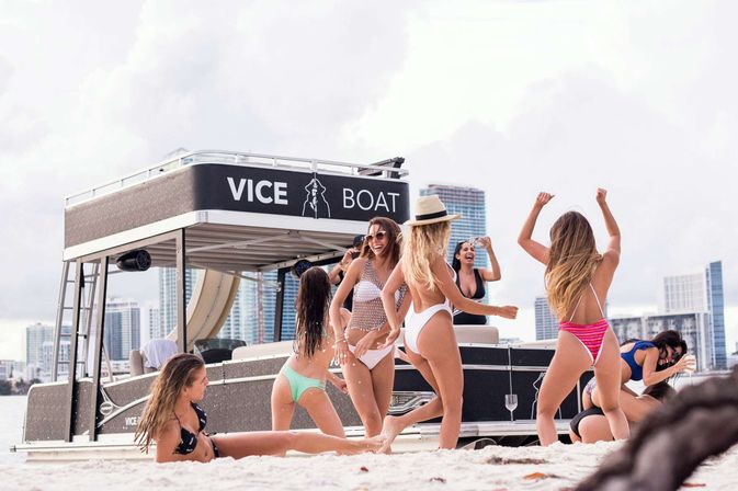 Beach boat party: a group of women in swimsuits dancing and laughing on sand beside a party pontoon boat, with a coastal city skyline of high-rise buildings in the background.
