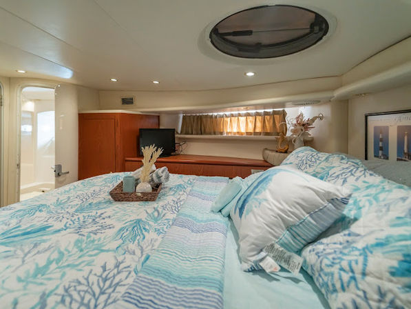 Cozy yacht cabin bedroom with coral-print bedding, round overhead porthole, wood cabinetry, small TV and decorative tray for a bright coastal marine interior