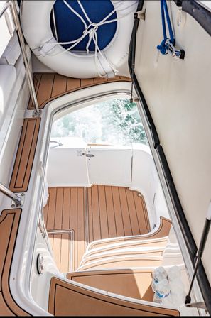 Overhead view of a sunlit motorboat deck with teak-style flooring, white life ring and rope, open transom revealing churning green sea, molded steps and bottled water on the swim platform.