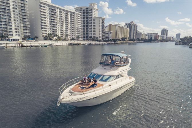Luxury motor yacht cruising a calm urban waterway past waterfront high-rise condominiums on a sunny day, with people relaxing on the bow