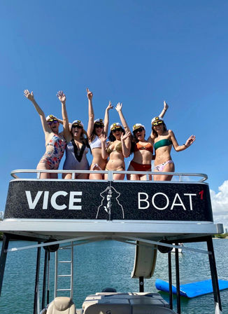 Six friends in bikinis and captain hats with arms raised, cheering on the upper deck of a pontoon party boat over a sunny lake under a clear blue sky