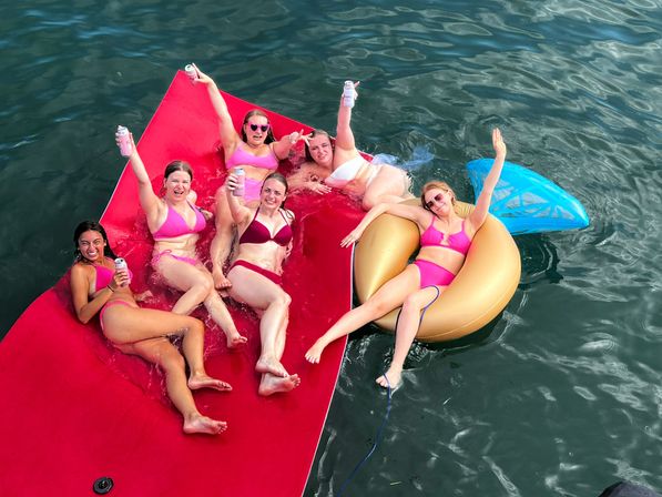 Six friends in pink and red bikinis lounging and cheering with canned drinks on a large red floating mat and a gold donut tube in green lake water — summer float party.