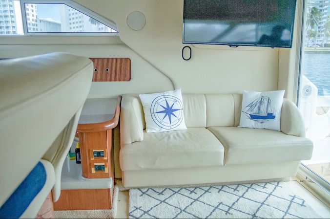 Yacht interior salon with beige leather sofa, nautical compass and sailboat throw pillows, wall-mounted TV, wood accents and water view through sliding glass door