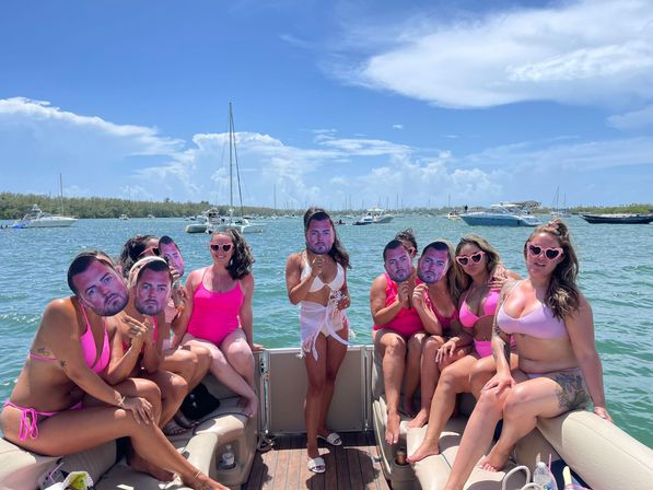Group of women in pink swimsuits enjoying a pontoon boat party in a sunny bay, some wearing heart-shaped sunglasses and holding oversized cutout masks of a man's face, with anchored sailboats and blue sky in the background.