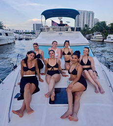 Seven women in black swimsuits relaxing on the bow of a white yacht at a waterfront marina with palm trees, high‑rise skyline, and an American flag