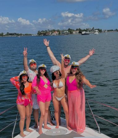 Six friends in captain hats and vibrant swimwear posing and cheering on the bow of a yacht in a sunny coastal bay with waterfront homes