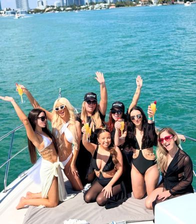 Seven women in swimsuits cheering with mimosas on a sunny yacht over turquoise water, coastal city skyline visible on the horizon.