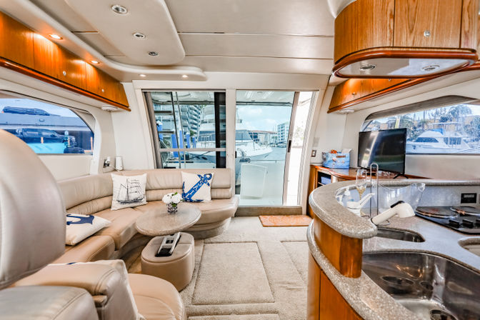 Sunlit luxury yacht interior salon and galley with curved beige leather seating, nautical throw pillows, small coffee table, curved stone countertop and sink, flat-screen TV, and sliding glass doors opening to a marina view.