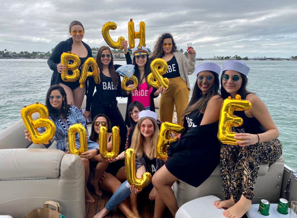 Group of women on a boat celebrating a bachelorette party, wearing sunglasses and sailor hats, bride in pink and a captain hat, holding shiny gold balloon letters with calm coastal water and cloudy sky in the background.