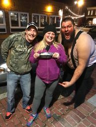 Three people posing on a brick sidewalk at night on a downtown main street — a woman in a purple hoodie holding a sticker, a man in camo on the left, and a roaring costumed performer with fake blood and suspenders on the right under string lights