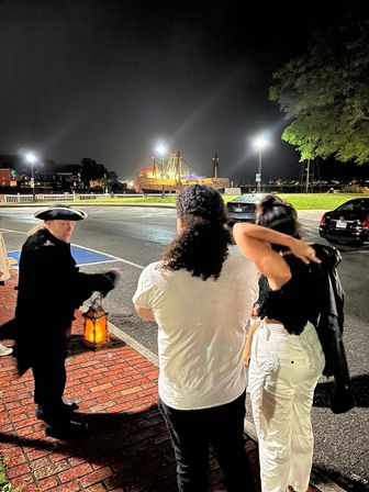 Lantern-lit waterfront at night: a costumed guide in colonial attire talks to two visitors on a brick sidewalk, with a brightly lit historic ship moored in the harbor under streetlights.