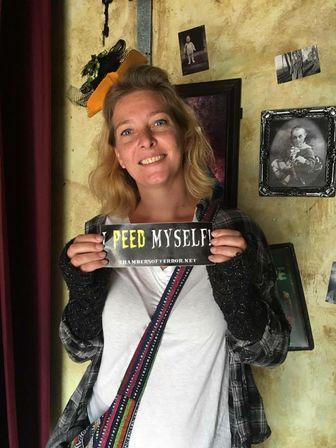 Smiling woman wearing a tiny witch hat and plaid shirt holds a novelty “I peed myself” sign in a spooky haunted-attraction photo booth with vintage black-and-white portraits on a textured yellow wall.