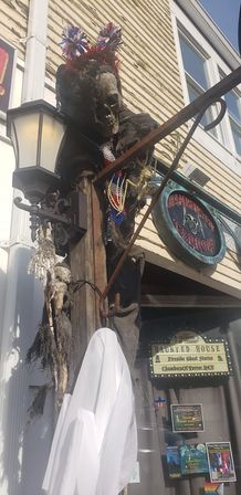 Spooky haunted-house storefront Halloween display: weathered skull in a tattered black cloak with red-white-blue pom-pom headband on a lamp post, hanging white ghost sheet and eerie sign on a clapboard building.