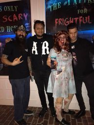 Group of four posing outside a haunted house at night — one in a bloodied vintage nurse costume holding a haunted attraction flyer, friends in dark casual clothes, brick sidewalk and spooky “SCARY”/“FRIGHTFUL” signage behind them