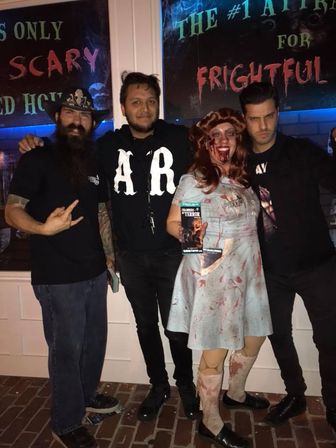 Group of four posing outside a haunted house at night — one in a bloodied vintage nurse costume holding a haunted attraction flyer, friends in dark casual clothes, brick sidewalk and spooky “SCARY”/“FRIGHTFUL” signage behind them