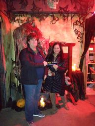 Two people pose in an indoor Halloween haunted-house photo-op surrounded by cobwebs, skull, flickering candles, a pumpkin and a small sign reading "I peed myself".