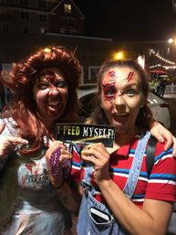 Two women in zombie makeup and torn costumes on a lit city street at night, smiling and holding a humorous “I peed myself!” sign at a haunted-event