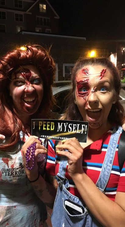 Two women in zombie makeup and torn costumes on a lit city street at night, smiling and holding a humorous “I peed myself!” sign at a haunted-event