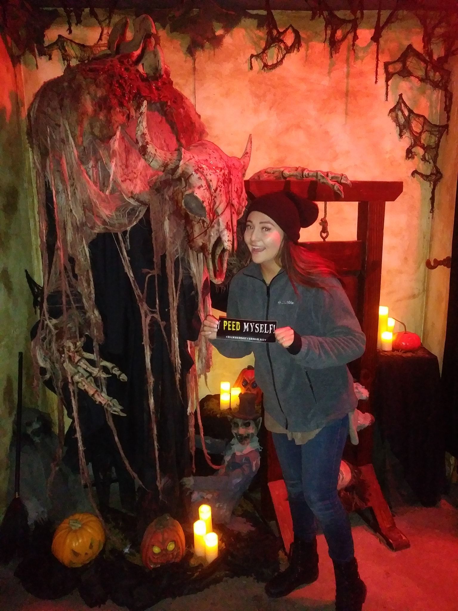 Person posing at an indoor haunted-house attraction with a giant horned skull draped in tattered cloth, glowing jack-o'-lanterns, flickering LED candles and a wooden guillotine prop, holding a humorous 'Peed Myself' sign.