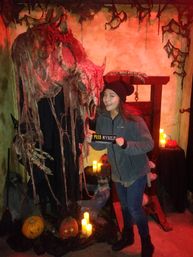 Person posing at an indoor haunted-house attraction with a giant horned skull draped in tattered cloth, glowing jack-o'-lanterns, flickering LED candles and a wooden guillotine prop, holding a humorous 'Peed Myself' sign.