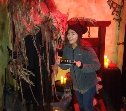Person posing at an indoor haunted-house attraction with a giant horned skull draped in tattered cloth, glowing jack-o'-lanterns, flickering LED candles and a wooden guillotine prop, holding a humorous 'Peed Myself' sign.
