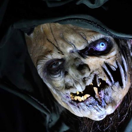 Close-up of a hooded, decayed witch-like horror prop with a glowing blue eye, yellow rotting teeth and torn mouth, spooky Halloween haunted-house decoration.