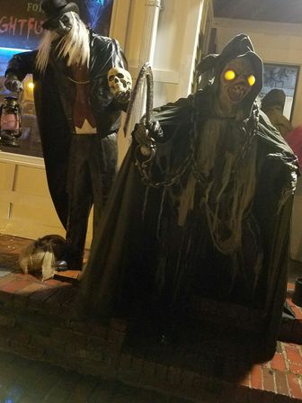 Halloween storefront display at night featuring two life-size spooky figures: a hooded reaper with glowing yellow eyes and chains, and a tall top-hatted figure holding a skull and lantern on brick steps.