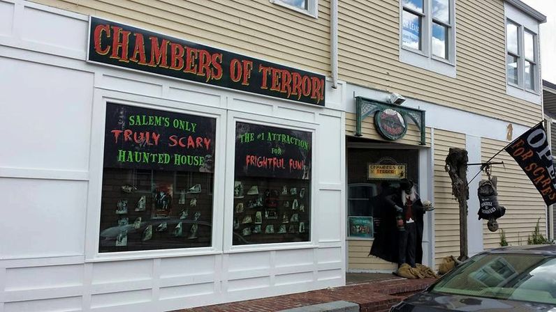 Spooky Salem haunted attraction storefront with 'truly scary' window signs, a costumed mannequin vampire at the doorway and a hanging skeleton prop on a wood‑sided building.