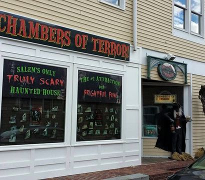 Spooky Salem haunted attraction storefront with 'truly scary' window signs, a costumed mannequin vampire at the doorway and a hanging skeleton prop on a wood‑sided building.