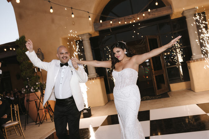 Bride in a strapless beaded white gown and groom in a white tux jacket dance on a black-and-white checkered floor outside a venue, arms outstretched amid sparklers and string lights.