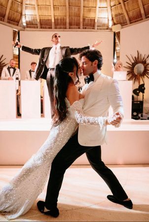 Bride and groom share a dramatic dip-and-kiss first dance under a thatched palapa roof, the bride in a beaded lace gown and the groom in a white tux while a live band and singer perform on stage behind them.