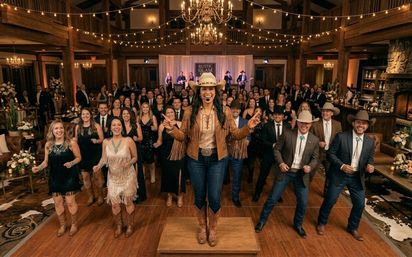 Country wedding reception in a rustic barn venue under string lights and chandeliers, a smiling line-dance leader in a cowboy hat, fringed jacket and boots on a small platform leads a large crowd of guests in western attire and evening dresses on the wooden dance floor