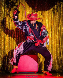 Stage performer in a red sequined cowboy hat and fringed black costume cracking a red whip in front of a glittering gold curtain